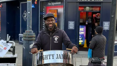 A street musician poses with drumsticks raised by a steel drum with Mighty Jamma written on it. He is smiling and wearing a black jacket with his stage name on it a black hoodie and black cap. A betting shop can be see behind him and a shopper walking past.