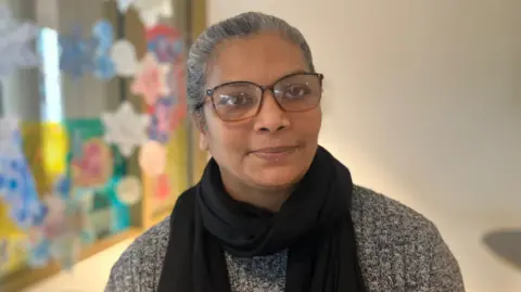 BBC/Elizabeth Baines A woman with grey hair and glasses looks at the camera. She wears a black scarf and a grey jumper.