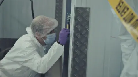 A forensic officer wearing a white hazard outfit and hairnet, purple rubber gloves and a clinical face mask, kneels down next to a storage locker. They are gripping a padlock that is connected to a metal bolt. To one side is yellow "Do not enter" police tape.