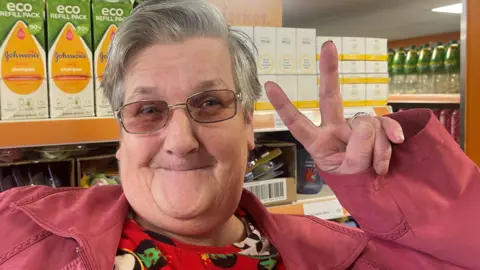 BBC An elderly woman with glasses makes a 'peace' sign in front of the supermarket shelves