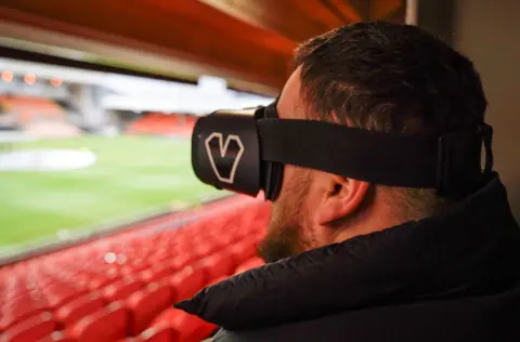 Jon Attenborough, a man with short dark hair and a beard, looks over an empty football stadium while wearing a headset that covers his eyes