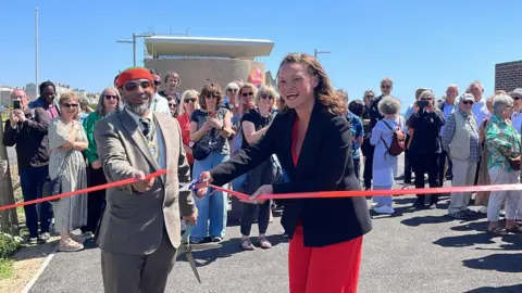 George Carden/BBC Bella Sankey cutting the red ribbon on Hove seafront with a crowd of people behind her. She is wearing a black blazer and red dress and smiling. Next to her is mayor Mohammed Asaduzzaman wearing a brown suit and red and green hat and sunglasses with his civic chain on