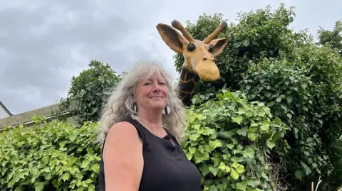 Valerie Boakes stands beside Julian the giraffe before he was damaged. She has long grey hair and a is wearing a black top, and is standing outdoors in front of some dense green foliage. Rising behind the greenery is a tall, lifelike giraffe sculpture. Its patterned neck and gentle face lean out of the greenery as if peering out.
