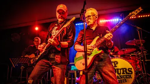 Two men on stage, Steve Young and Nik Kershaw. They are both holding guitars. In the background is another guitarist and a set of drums.