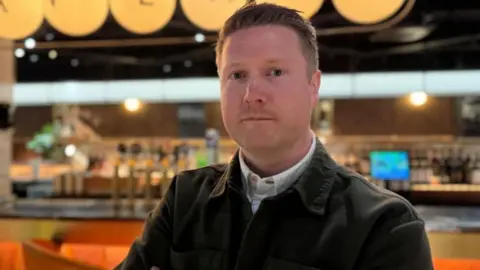 Paul Anderson, a man with short, brown hair looks into the camera. He is wearing a white shirt and a brown buttoned up jacket. He is standing in front of a bar. The background of the image is blurred.