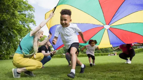 Children running underneath a multicoloured parachute being held up by grown ups on a field