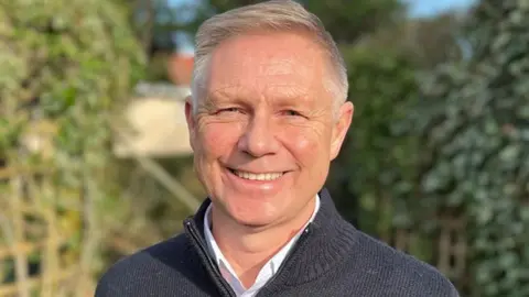 Head and shoulders image of David Morris smiling in a garden in the sunshine. He has short white hair with a side parting and is wearing a white shirt under a dark zip up jumper with a stand up collar