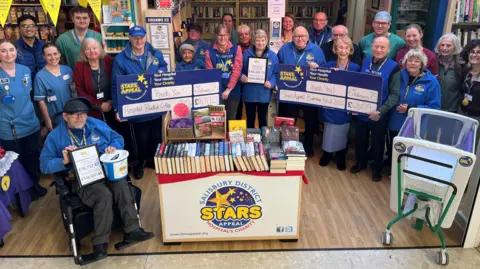 A large group of Stars Appeal volunteers are gathered around a book stand in the charity's shop. They hold giant cheques representing the money they have raised. One volunteer holds a collecting bucket. An empty baby incubator can be seen on one side.  