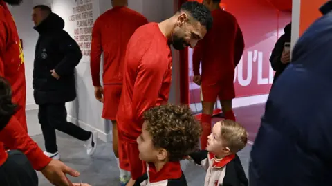 Family photo Mo Salah, in a red training top and shorts, shakes hands with a young boy who has light brown hair and is wearing a white and black zip up jacket.