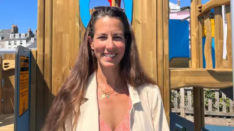 A picture of Emily Jennings standing in front of some play equiment. She is wearing a pink shirt and a white blazer. She has long brown hair.
