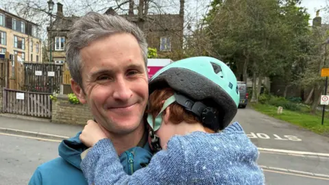 A man stands outside with a stone building in the background. There are trees behind him. He has grey hair and is wearing a blue hooded jacket. He is smiling into the camera and holding a child in his arms. The child is wearing a blue cycle helmet and a blue knitted jumper and his face is turned away from the camera and buried in the man's shoulder.