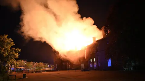 STUART COWPER A large fire at a former primary school in Perth. The fire is bright orange and can be seen coming out of the top of the building, which is dark. The sky is also dark.