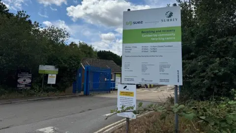A sign says 'Recycling and recovery, Camberley community recycling centre,' The entrance to the centre can be seen in the background.