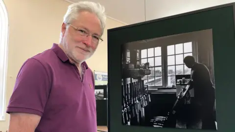 Wherry Lines Community Rail Partnership David Pearce smiles at the camera while standing next to a black and white image of a man standing in a railway operator room. David has white grey hair and wears a burgundy polo T-shirt. 