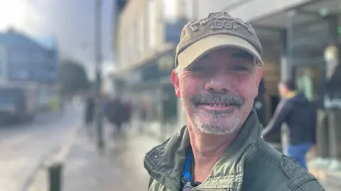 A man with grey goatee beard, green baseball cap and army jacket.