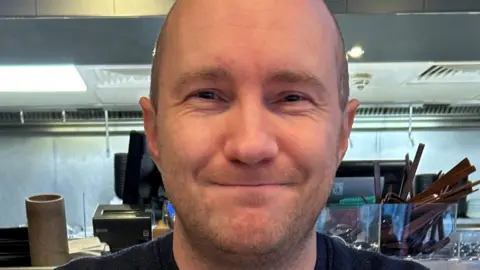 A bald man with short stubble smiles in a restaurant or café, wearing a dark sweater.