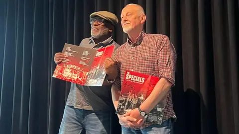 Universal Music Group Lynval Golding, wearing a flat cap and glasses, holds up a copy of the new album as he stands in front of a dark curtain. Horace Panter is standing beside him wearing a red checked shirt with a copy under his arm. 