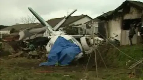 A damaged plane with a blue tarpaulin on its nose. You can see fire damage to the house on the right hand side.