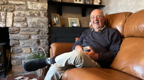 John Sibley, wearing a black jumper and beige chinos, sits on his leather sofa smiling. He is wearing glasses and slippers, with a cup of coffee in his hand. There are family photos and a fireplace in the background.