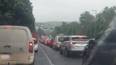 Una larga fila de coches se extiende a lo largo de dos carriles de tráfico. El cielo está oscuro y las carreteras mojadas.