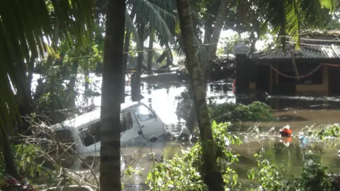 Sian Hughes A white van is partly submerged in water, surrounded by palm trees and flooded huts