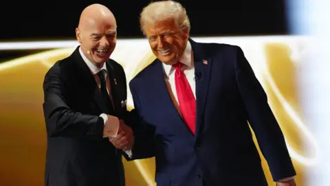 Donald Trump is shaking heads with Gianni Infantino the President of Fifa. They are both on stage wearing dark suits. Donald is wearing a red tie and a badge of the American flag.