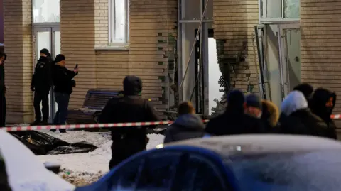 Scene of the explosion in which Igor Kirillov was reported killed. A damaged wall shows blackened and missing bricks, while two armed guards stand to the left and a crowd looks on from behind a red-and-white striped security cordon.