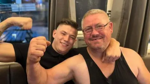 Handout A father and son posing with their right arms showing their muscles. They're smiling at the camera and sat down on a leather sofa