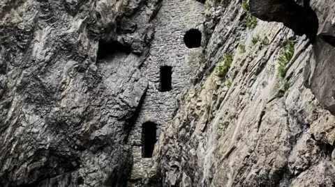 Wide view looking up through a rocky gorge with jagged grey cliff walls on both sides. A tall, narrow stone structure is embedded between the cliffs, with small window openings, and a dry, pebble-covered riverbed lies at the bottom.