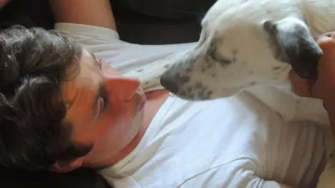 Lesley Wade Aaron Armstrong pictured with a dog. The dog, which is white with black spots, is laying on Aaron's chest. Aaron is pulling a kissing face at the dog, and is wearing a white t-shirt.