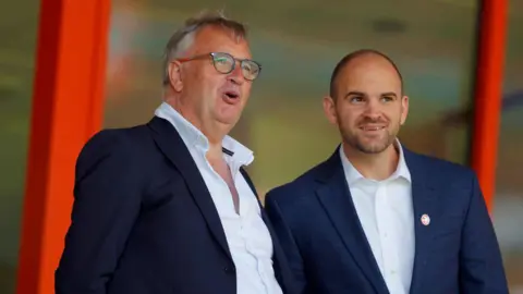 Walsall co-chair Ben Boycott stands on the right in a navy suit in the stands at the Bescot Stadium