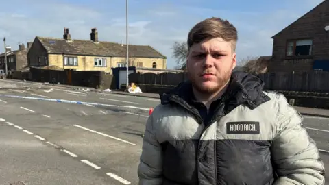 Heidi Tomlinson/BBC A man with a grey and black puffer coat stands at the side of a road. Behind him is police tape around the scene