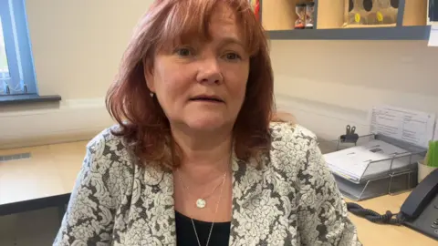 A women in a black and white floral blazer sits in an office, she is wearing to small silver necklaces 