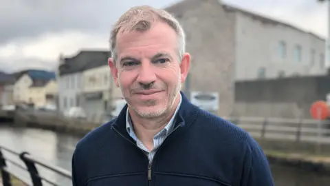 A man standing looking at the camera with a slight smile. Behind it is a canal and some buildings.