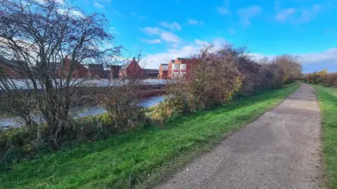 The location of the proposed bridge. A wide canal towpath is seen on this side of the canal, with an under-construction housing estate on the other side. Some trees are seen just on this side of the canal.