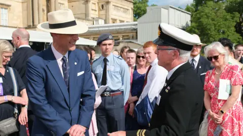 The Duke of Edinburgh’s Award The Duke of Edinburgh wears a sun hat and blue suit as he smiles and speaks to a man in a military-style uniform. A small smartly-dressed crowd is behind them.