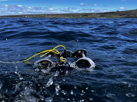 A small underwater drone sits on the surface of the sea, attached by a yellow cable.  The land is seen in the distance.