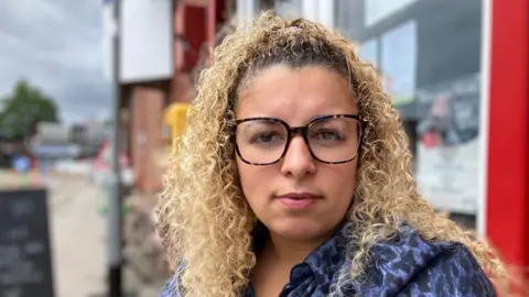 A woman with large square spectacles looks at the camera. She has curly blonde hair and a blue shirt on. Behind her, a cafe is blurred.