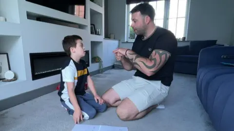 A boy and a man are sat on a floor in a living room. The man has dark-brown short hair and is wearing cream shorts and a black T-shirt. The boy has dark-brown hair and is wearing a blac, white and yellow T-shirt and grey and white track suit bottoms. The boy looks at the man has he uses his two hands to demonstrate a symbol.