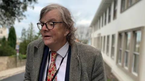 BBC A man wearing glasses with medium length grey hair, wearing a brown jacket, purple shirt and a red patterned tie.