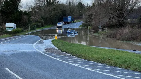 A blue Mini car floats in floodwater on a main road through the Cotswolds. Out of the water, on the road behind, there is a police van and a lorry, and a motorhome can be seen on a grass verge. 