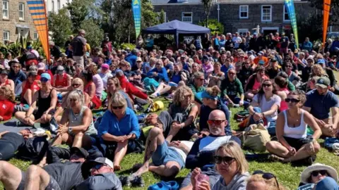 A crowd of people sitting down on grass looking at a big screen