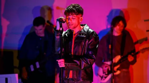 Getty Images Louis Tomlinson wears a black leather jacket as he performs in front of two guitarists on a stage bathed in the glow of red and purple stage lights. His eyes are closed as he sings, one hand resting on the microphone in front of him and the other tightly gripping the stand below it.
