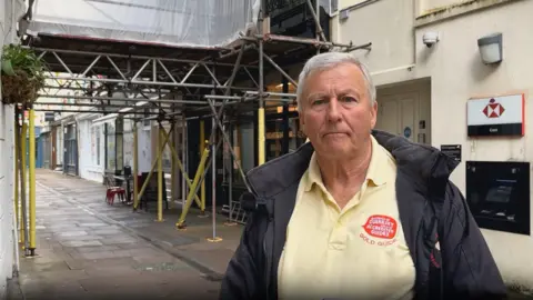 BBC The image shows the chair of the Guild of Guides, Ken Wheeler standing on a street. He has short grey hair and is wearing a yellow polo shirt and a black jacket. Behind him is scaffolding attached to a building. 