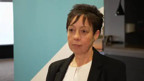 Dr Lemmens has short, dark hair spiked up at the back. She wears a black blazer over a white blouse