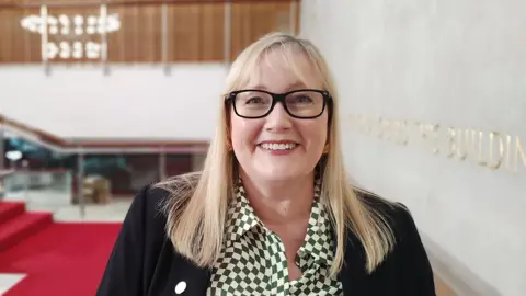 Newcastle City Council Karen Kilgour smiling at the camera. She has medium-length straight blonde hair with a fringe. She is wearing black-rimmed glasses, a green-and-white patterned shirt and a black suit jacket. 