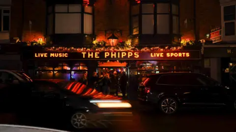 The Phipps The exterior of a pub in the dark with "The Phipps" illuminated in orange lights with people standing outside and cars driving past.