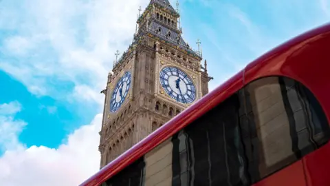 Getty Images Big Ben clock tower at the Palace of Westminster in London viewed from below, with a red double‑decker bus passing in the foreground against a blue sky with scattered clouds.