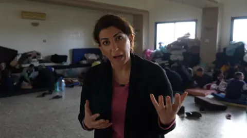 BBC Beirut correspondent Carine Torbey speaks to the camera in a room with displaced Alawites