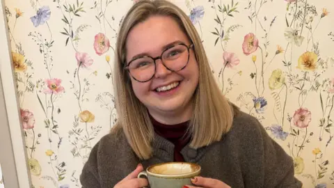 Evie Brooks A blonde haired woman with tortoise shell glasses sitting smiling at the camera holding a mug of coffee. Behind her is floral wallpaper and she is wearing a brown knit cardigan. 
